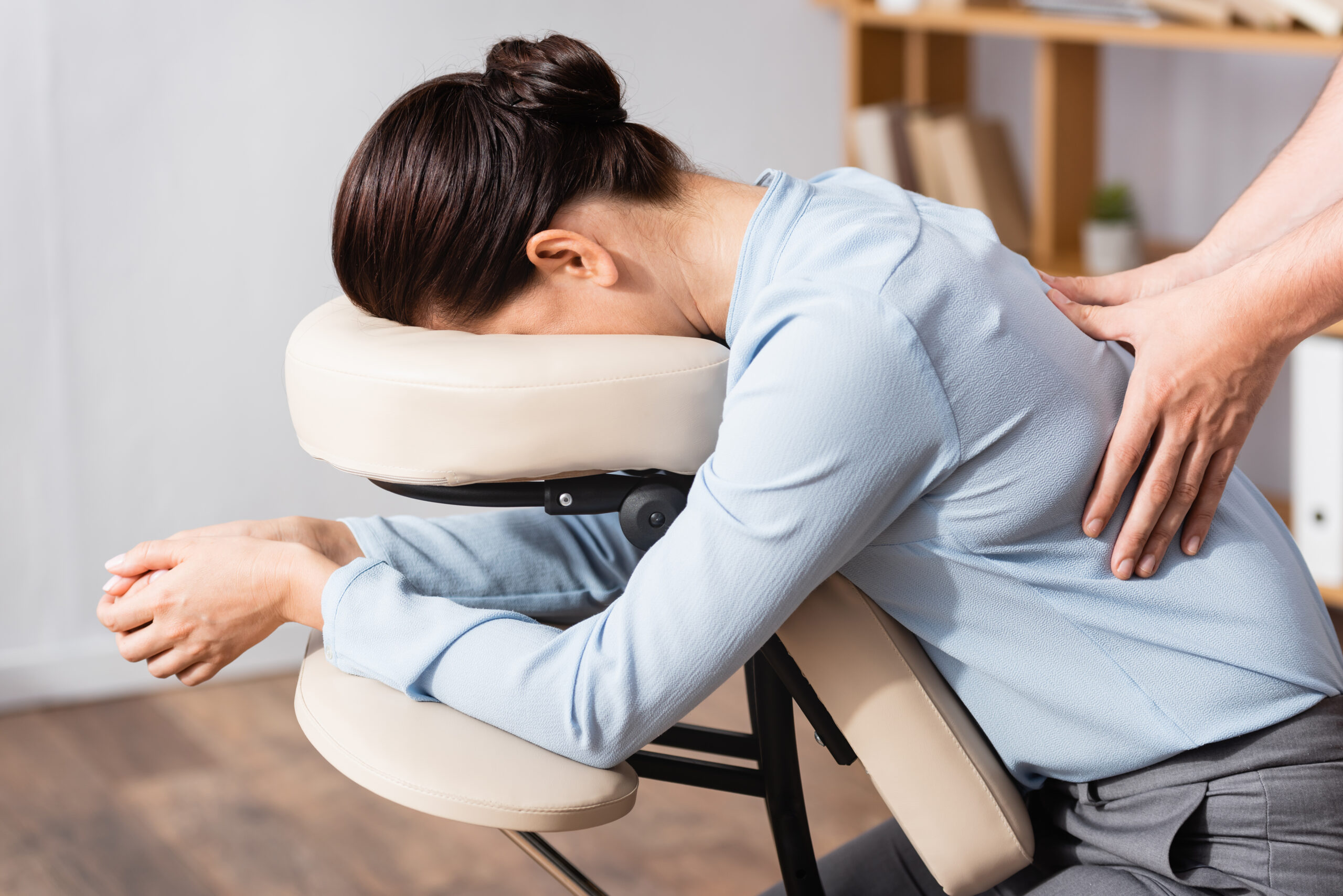 Side view of masseur doing seated massage of back for brunette businesswoman with blurred office on background